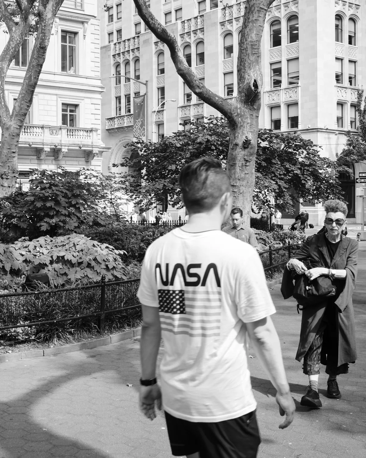 A figure in a NASA t-shirt with American flag graphic walks through Madison Square Park in the Flatiron District, their motion creating a gentle blur against the static urban tableau. An elderly man in dark coat and sunglasses sits contemplatively on a bench, framed by the ornate Beaux-Arts and Renaissance Revival architecture that defines this historic Manhattan neighborhood. The dappled shadows of mature plane trees create a natural canopy over the scene, while the classical facades of the surrounding buildings speak to New York's gilded age grandeur.