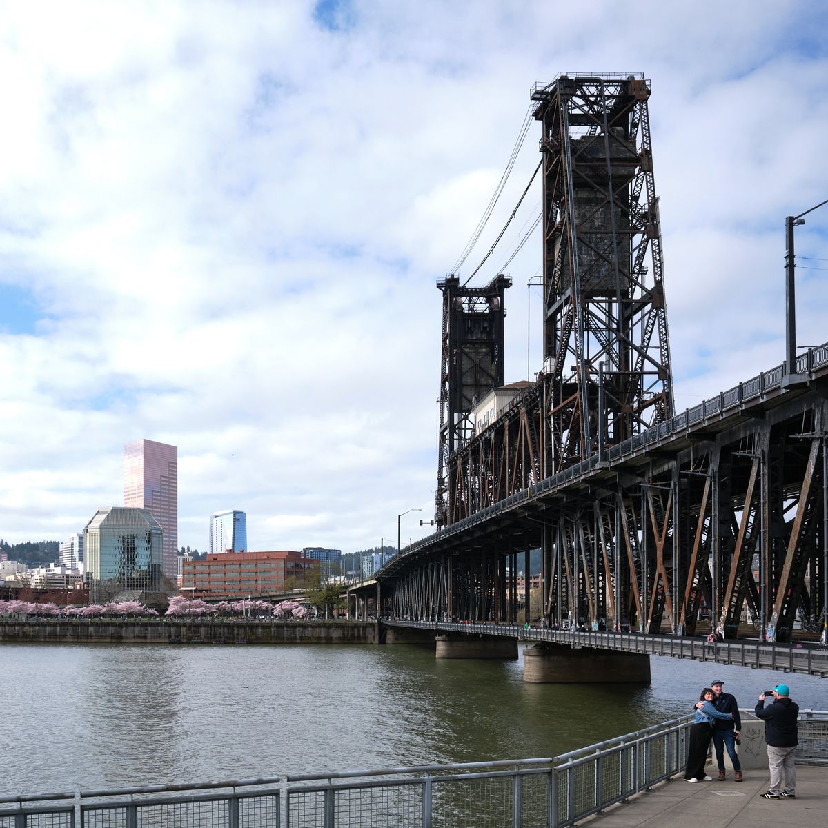 The weathered steel towers of Portland's Steel Bridge rise majestically against a soft spring sky, their industrial bones creating dramatic vertical lines above the Willamette River. In the foreground, visitors gather along the Eastbank Esplanade's waterfront promenade, while delicate cherry blossoms create a romantic pink haze near Tom McCall Waterfront Park on the distant shore. The scene captures the harmonious contrast between Portland's gritty industrial heritage and its celebrated urban greenspace, with the city's modern skyline providing a backdrop of glass and brick beneath clouds that filter the Pacific Northwest light into gentle, diffused tones.