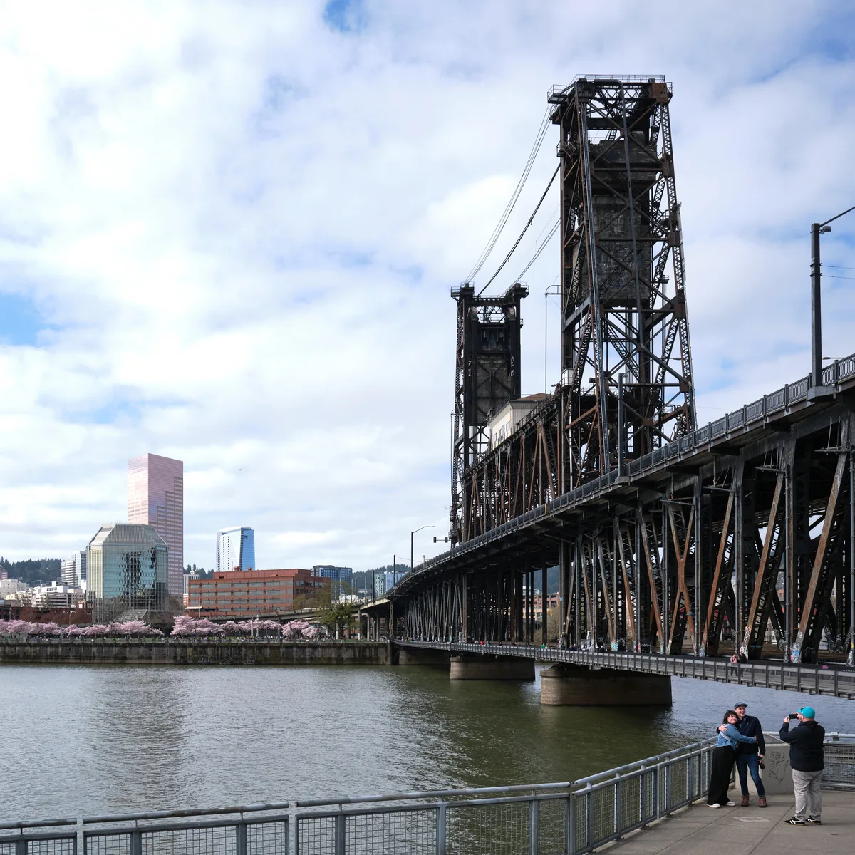 The weathered steel towers of Portland's Steel Bridge rise majestically against a soft spring sky, their industrial bones creating dramatic vertical lines above the Willamette River. In the foreground, visitors gather along the Eastbank Esplanade's waterfront promenade, while delicate cherry blossoms create a romantic pink haze near Tom McCall Waterfront Park on the distant shore. The scene captures the harmonious contrast between Portland's gritty industrial heritage and its celebrated urban greenspace, with the city's modern skyline providing a backdrop of glass and brick beneath clouds that filter the Pacific Northwest light into gentle, diffused tones.