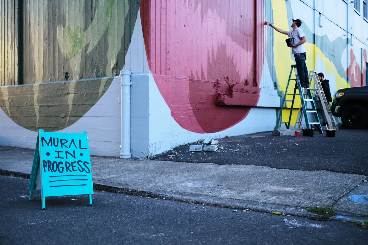 An artist perched on a ladder applies bold strokes to a sweeping geometric mural adorning the exterior wall of Coast Auto Supply on Southeast Stark Street. The composition features flowing bands of sage green, dusty rose, and sunshine yellow that curve across the weathered concrete surface. A cheerful turquoise sidewalk sign reading 'MURAL IN PROGRESS' sits on the cracked asphalt, while afternoon light catches the fresh paint and casts long shadows across the urban streetscape.