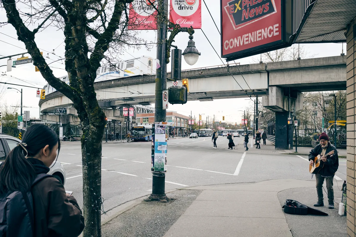 A street musician with an acoustic guitar performs beneath the weathered concrete spans of the North Grandview Highway overpass in Vancouver's Grandview-Woodland neighborhood. The moody overcast sky casts soft, diffused light over the urban intersection, where pedestrians cross the wide street and a hooded figure in the foreground observes the scene. Red convenience store signage and bare winter trees frame this intimate moment of street performance against the backdrop of Vancouver's transit infrastructure.