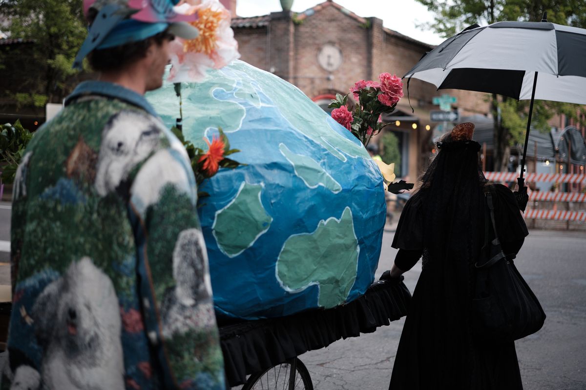 A street performance scene in Portland's Central Eastside district featuring performers with a large painted Earth globe prop decorated with flowers, while a person in dark clothing holds an umbrella nearby. The urban setting includes brick buildings and trees in the background.