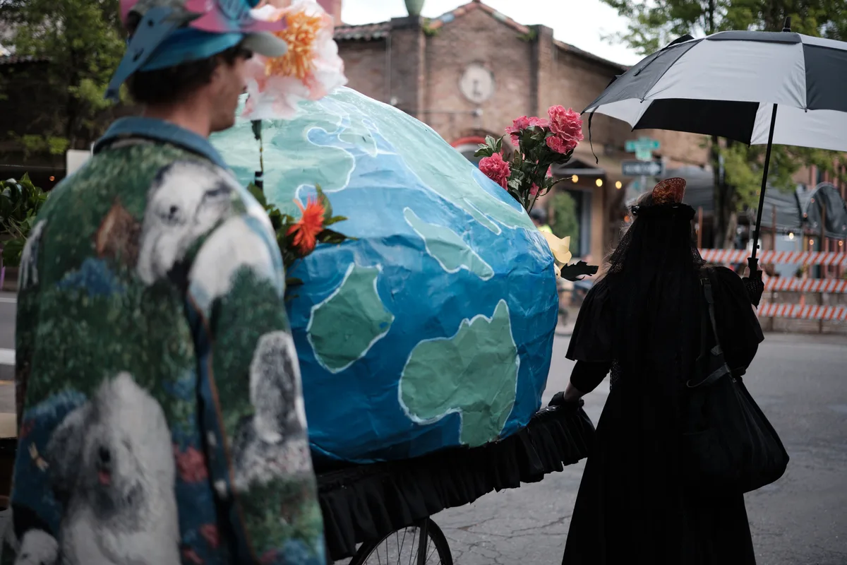 A street performance scene in Portland's Central Eastside district featuring performers with a large painted Earth globe prop decorated with flowers, while a person in dark clothing holds an umbrella nearby. The urban setting includes brick buildings and trees in the background.