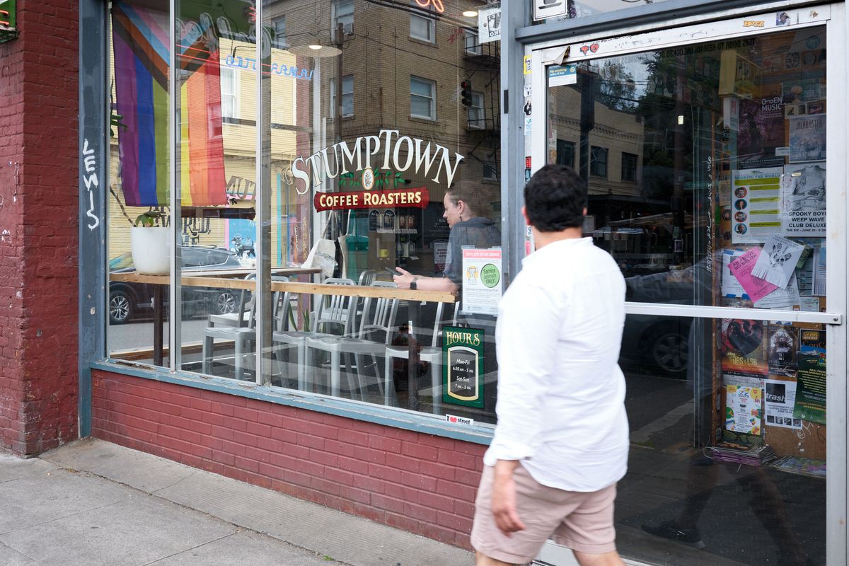A person in a white shirt walks past the storefront window of Stumptown Coffee Roasters on Southeast Belmont Street in Portland's Sunnyside neighborhood. The large windows display the coffee shop's interior with customers seated at tables, while colorful flyers and community notices are posted on the glass.