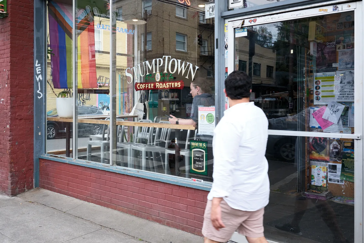 A person in a white shirt walks past the storefront window of Stumptown Coffee Roasters on Southeast Belmont Street in Portland's Sunnyside neighborhood. The large windows display the coffee shop's interior with customers seated at tables, while colorful flyers and community notices are posted on the glass.