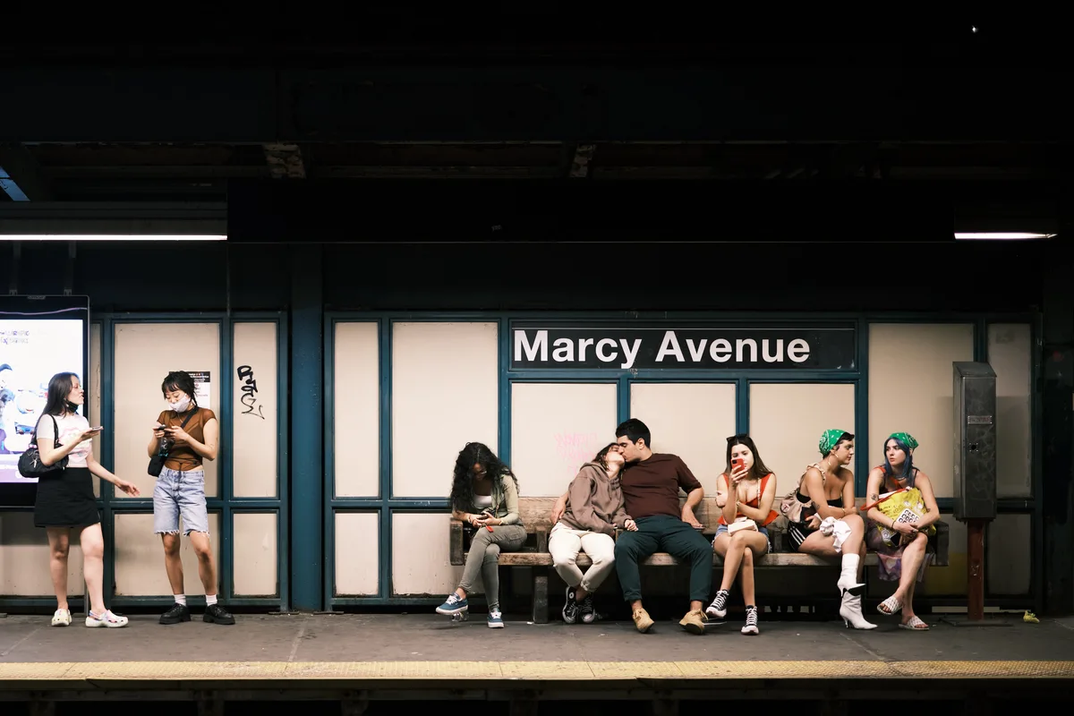 Passengers wait beneath the stark fluorescent lighting of Brooklyn's Marcy Avenue subway platform, their bodies creating a intimate tableau against the institutional green tiling. A couple shares a tender moment in the center of the bench while fellow commuters engage with their phones and conversations, each absorbed in their own urban rituals. The warm summer evening light filters through the station's windows, softening the harsh underground environment and illuminating this slice of contemporary New York transit life.