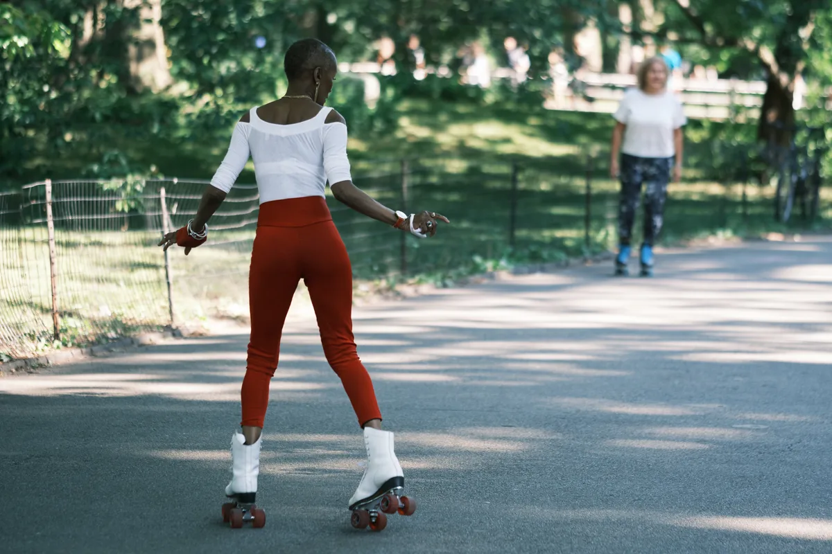 A woman glides gracefully on roller skates along a sun-dappled path in Central Park, her white off-shoulder top and vibrant red pants creating a striking silhouette against the verdant canopy. The golden afternoon light filters through the dense foliage, casting dancing shadows across the asphalt as park visitors enjoy the peaceful setting in the background. Her movement captures the effortless joy of urban recreation, while the shallow depth of field creates an intimate moment within the bustling heart of Manhattan.