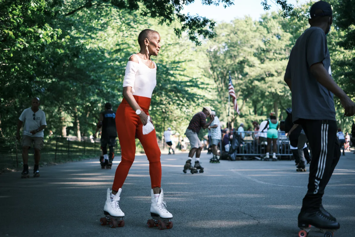 A woman in striking red pants and white off-shoulder top glides gracefully on white roller skates along Central Park's tree-lined pathways, her confident posture and gentle smile embodying the joy of summer recreation. The dappled sunlight filters through the lush green canopy above, creating a nostalgic atmosphere as fellow skaters and park-goers blur softly in the background. Her athletic stance and focused expression capture a moment of pure freedom and movement, while the classic roller derby aesthetic brings vintage charm to this contemporary urban scene.