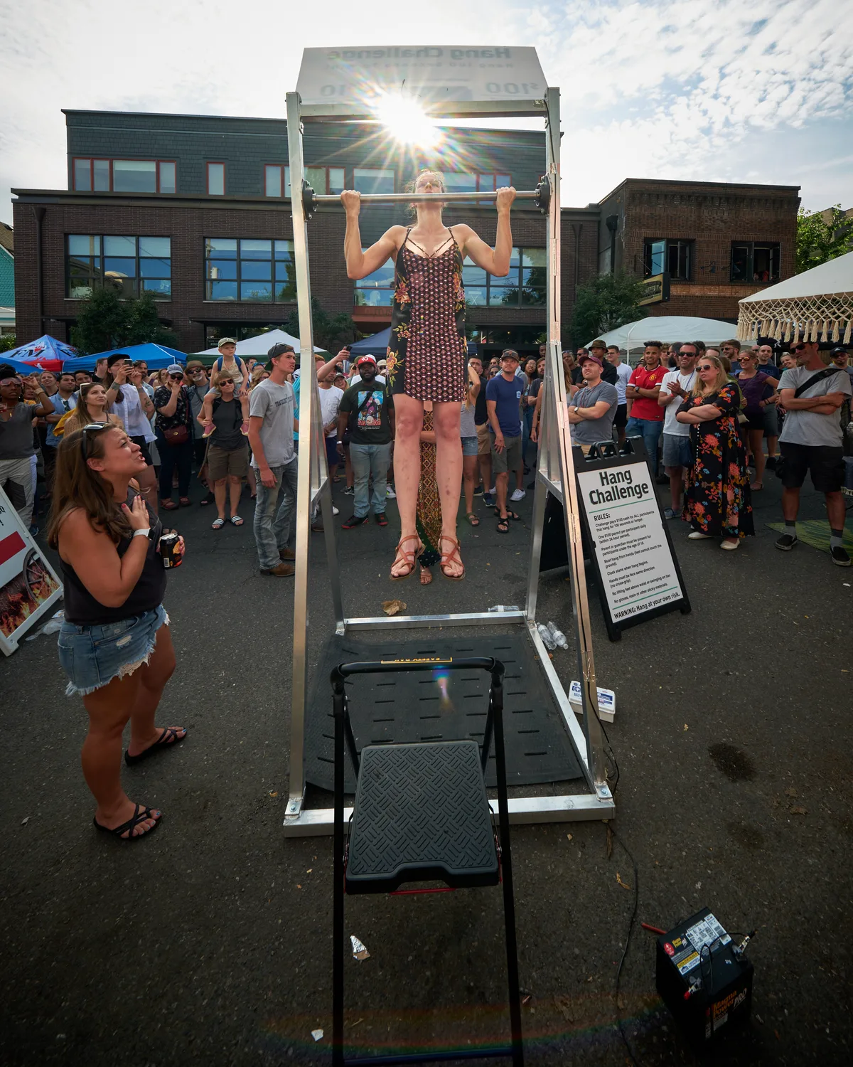 A determined woman in a geometric patterned dress executes a perfect dead hang from a pull-up bar at Portland's Mississippi Street Fair, her form silhouetted against the brilliant afternoon sun. The crowd of onlookers forms a semicircle around the Hang Challenge station, their faces a mixture of encouragement and awe as she demonstrates raw strength in an unexpected urban setting. The lens flare creates dramatic rays that pierce through the scene, while the contemporary brick architecture of North Mississippi Avenue provides a distinctly Portland backdrop to this moment of athletic determination.