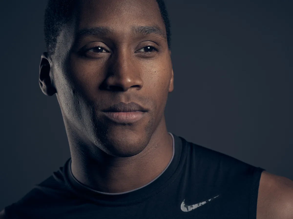 A close-up studio portrait of a young man wearing a black Nike sleeveless shirt, lit dramatically against a dark grey background. He gazes slightly off-camera with a calm, composed expression.
