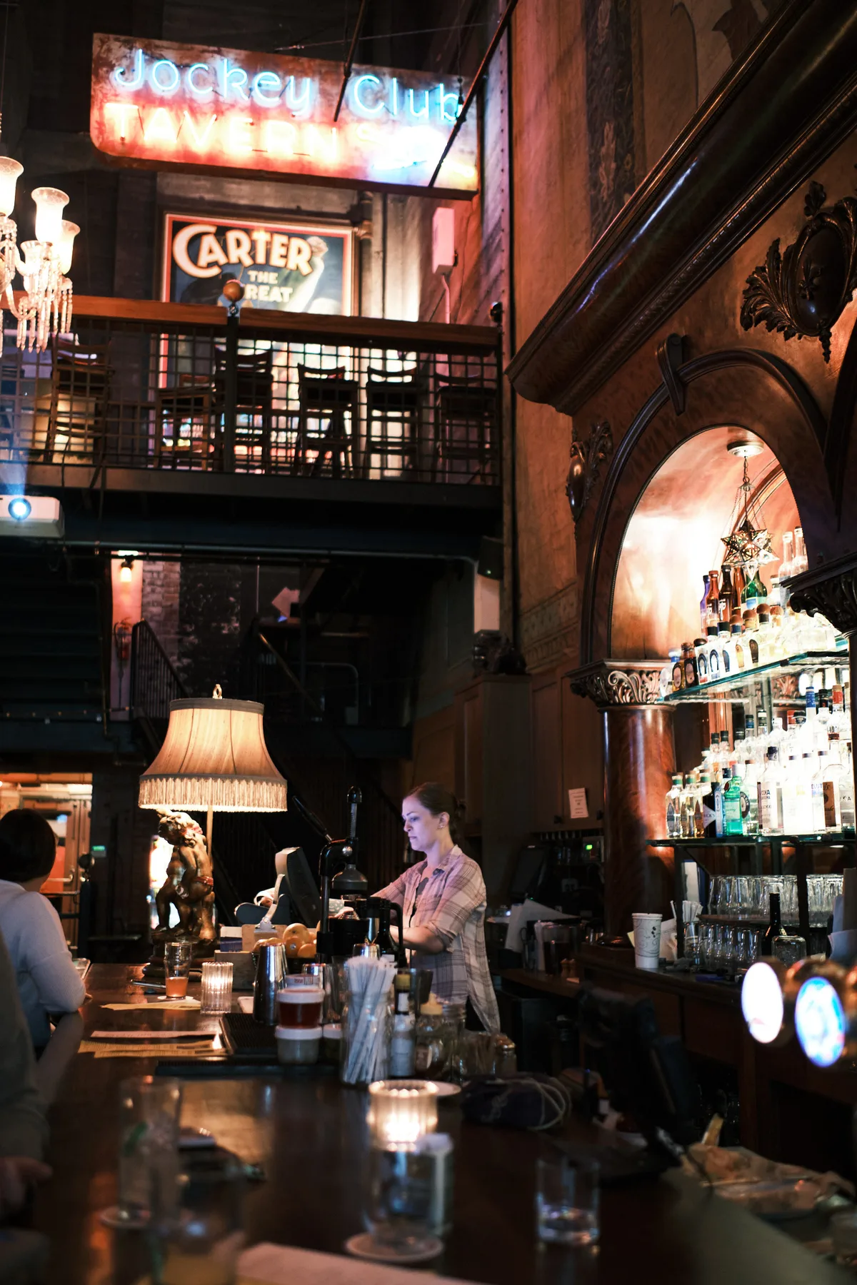 A bartender works behind the mahogany bar at Portland's historic Backstage Bar, illuminated by the warm glow of vintage neon signs reading 'Jockey Club' and 'Carter's The Great.' The atmospheric interior features soaring Gothic arches, ornate moldings, and a mezzanine level with iron railings, while fringed table lamps and carefully arranged bottles create theatrical shadows. The composition captures the establishment's vaudeville heritage through its dramatic lighting and layered architectural details.