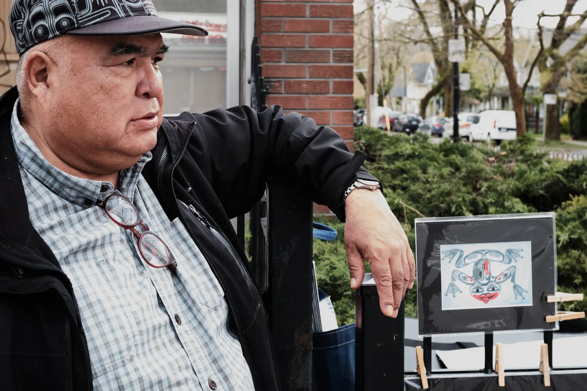 Brad Henry, a former firefighter turned traditional Tlingit artist, sits contemplatively beside his artwork on Commercial Drive in Vancouver's Grandview-Woodland neighborhood. The Indigenous artist, wearing a distinctive patterned cap and plaid shirt beneath a dark jacket, gestures toward a framed piece featuring traditional blue and red Tlingit designs. Natural daylight filters through the urban setting, highlighting the weathered brick architecture and spring foliage that frame this intimate portrait of cultural preservation and artistic dedication.