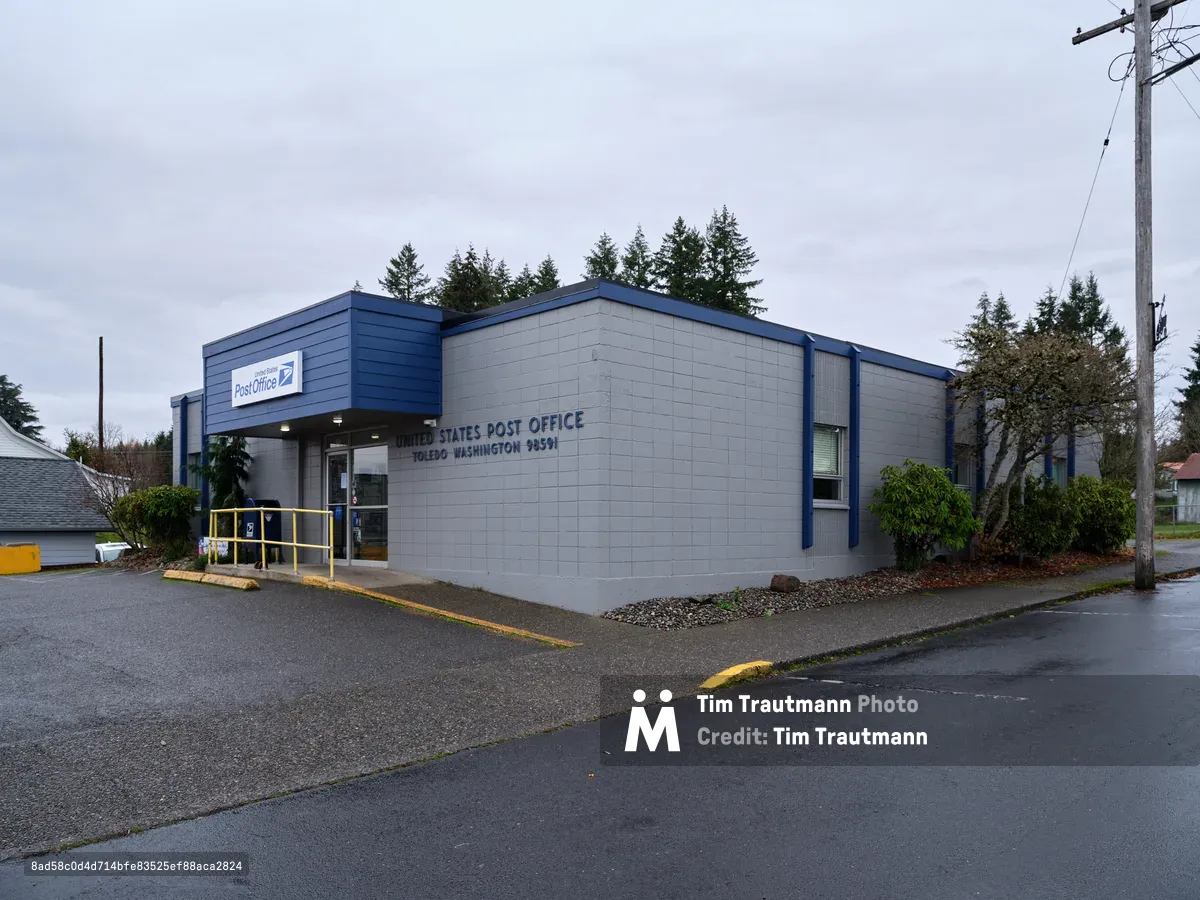 A modest single-story United States Post Office stands beneath the brooding gray skies characteristic of western Washington's maritime climate. The building's utilitarian design features white concrete block walls punctuated by blue trim, while towering evergreens create a dramatic backdrop against the overcast heavens. Wet pavement reflects the muted daylight, and yellow accessibility railings provide the only warm accent in this quintessentially Pacific Northwest municipal scene.