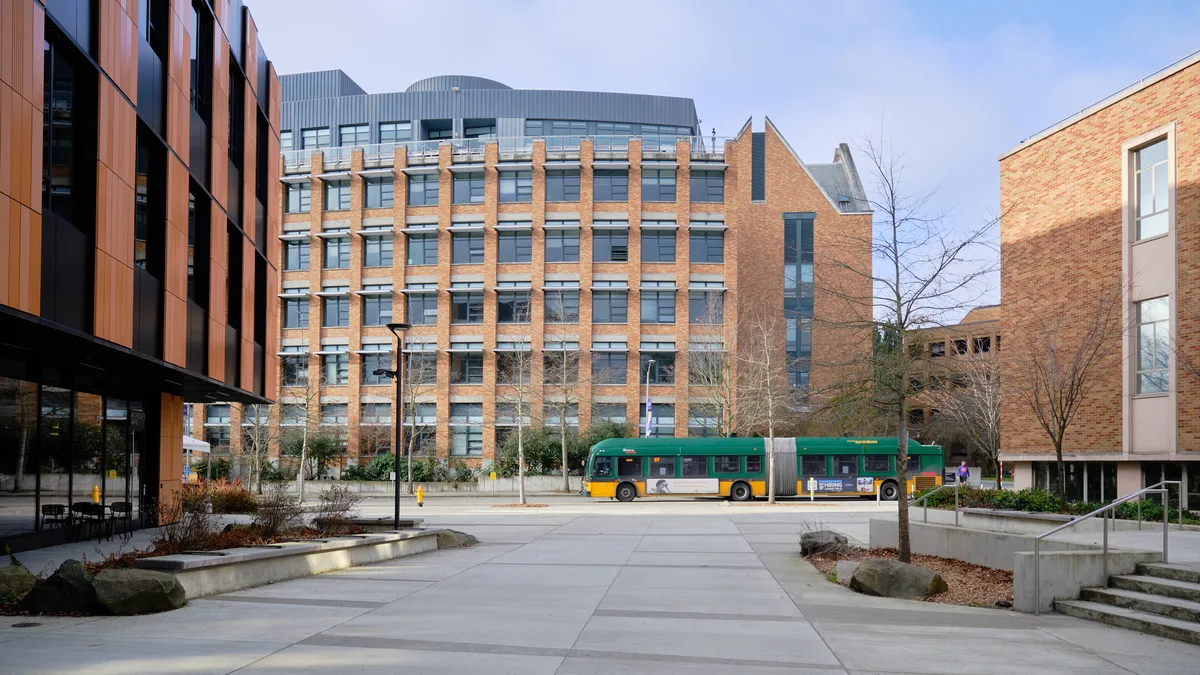 A King County Metro articulated bus glides through the University of Washington campus plaza, its green and gold livery creating a striking counterpoint to the warm brick facades of modern academic buildings. The late afternoon light casts gentle shadows across the concrete courtyard, where bare winter trees stand sentinel among carefully planned landscaping. The architectural composition frames the scene with geometric precision, balancing the institutional grandeur of the multi-story research buildings against the human scale of the transit corridor.