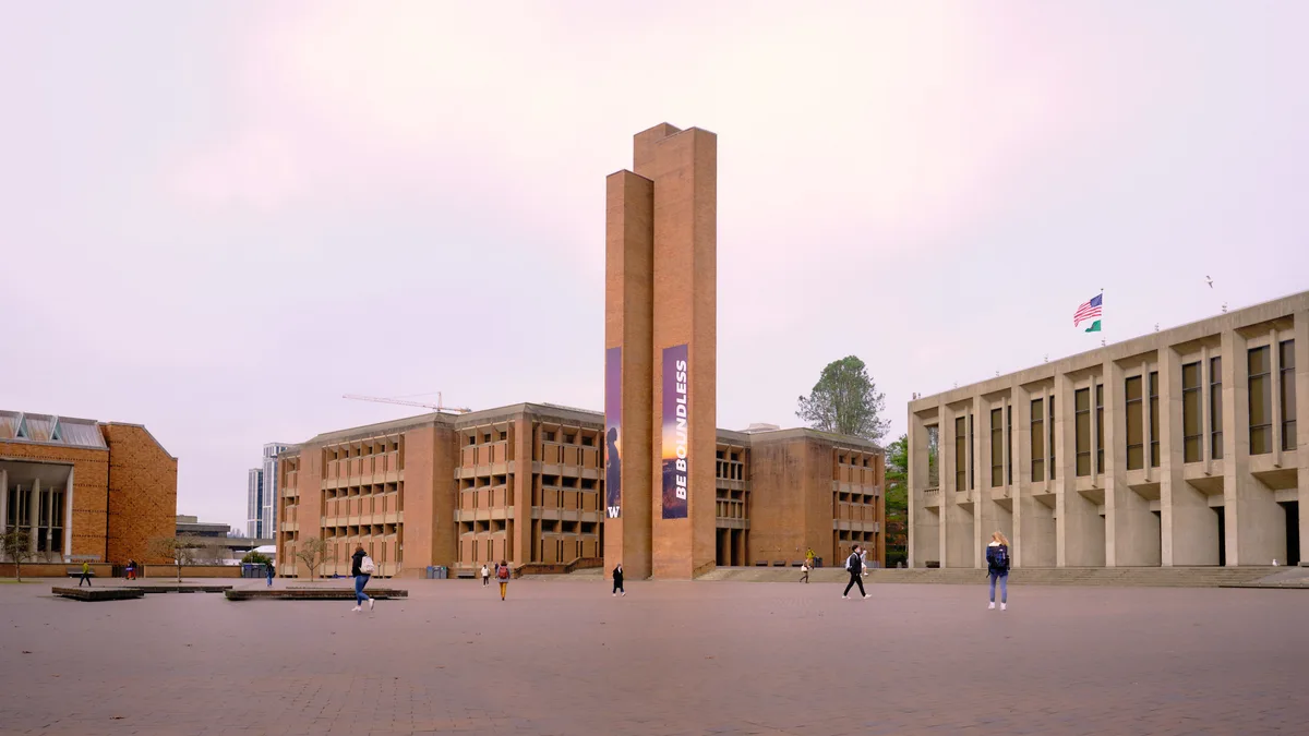The iconic Red Square at the University of Washington bathes in ethereal pink twilight, with Louis Kahn's brutalist Suzzallo Library tower standing sentinel against a pastel sky. Students traverse the vast brick plaza in scattered formation, their figures dwarfed by the monumental concrete and brick architecture that defines this celebrated campus heart. The geometric interplay between the library's vertical thrust and the horizontal expanse of surrounding academic buildings creates a striking study in modernist design, while the soft evening light transforms the utilitarian materials into something almost mystical.