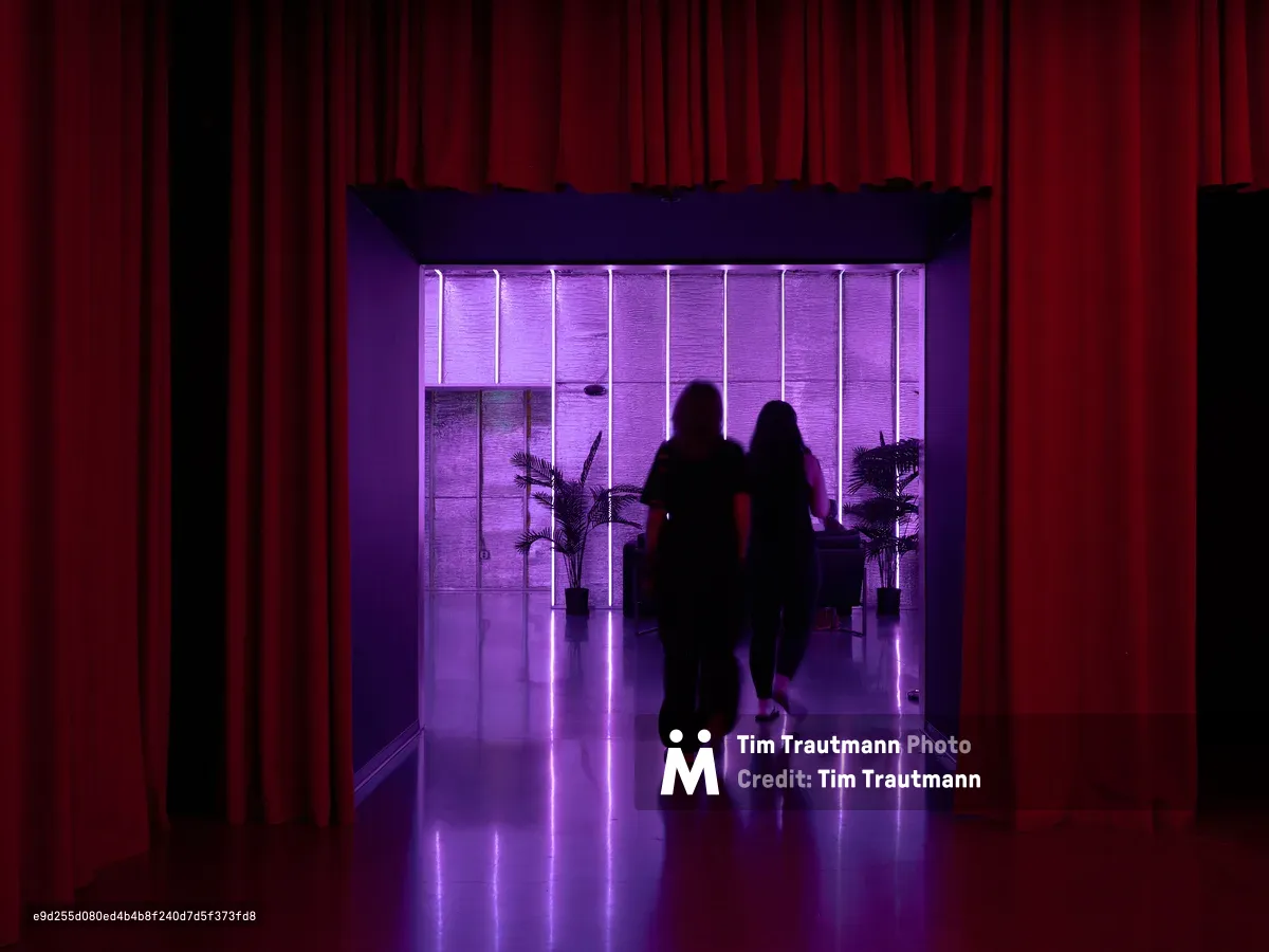 Two people walk as silhouettes through a dramatically lit modern interior corridor featuring purple neon lighting, red curtains, and reflective floors. The atmospheric lighting creates a striking contrast between the warm red fabric walls and cool purple illumination.