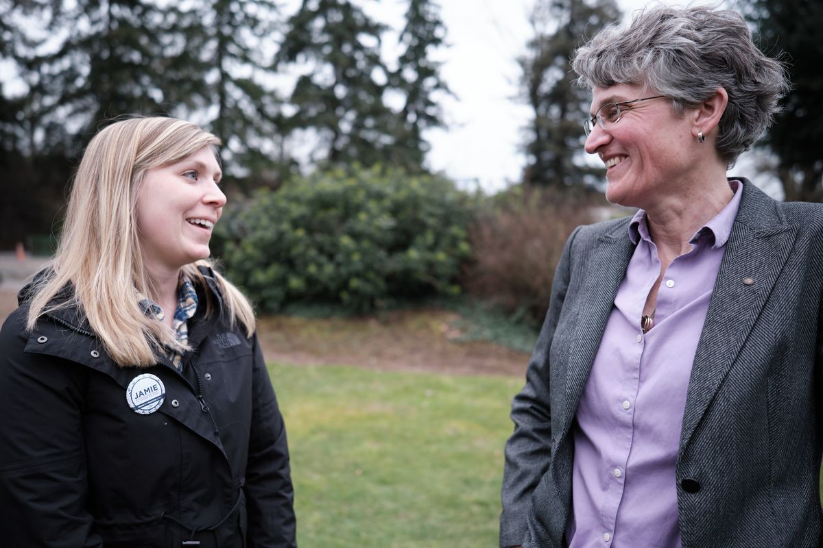 Two women stand facing each other in an outdoor park setting in Oregon City, Oregon, engaged in friendly conversation and smiling. The younger blonde woman on the left wears a dark jacket with a campaign button, while the older woman on the right wears glasses, a gray blazer, and purple shirt.