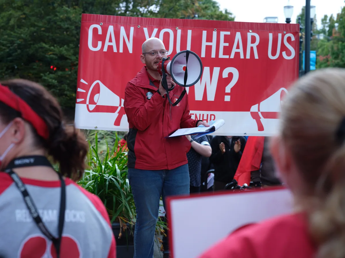A bald man in a vibrant red jacket commands attention as he speaks through a megaphone at a Kaiser Permanente nurses' strike rally in Portland's Lloyd District. Behind him, a bold red and white banner poses the rhetorical question 'CAN YOU HEAR US NOW?' - its message emphasized by megaphone graphics. The speaker holds papers in one hand while addressing a crowd of supporters wearing matching red and white strike colors, creating a sea of solidarity that fills the foreground with purposeful energy.
