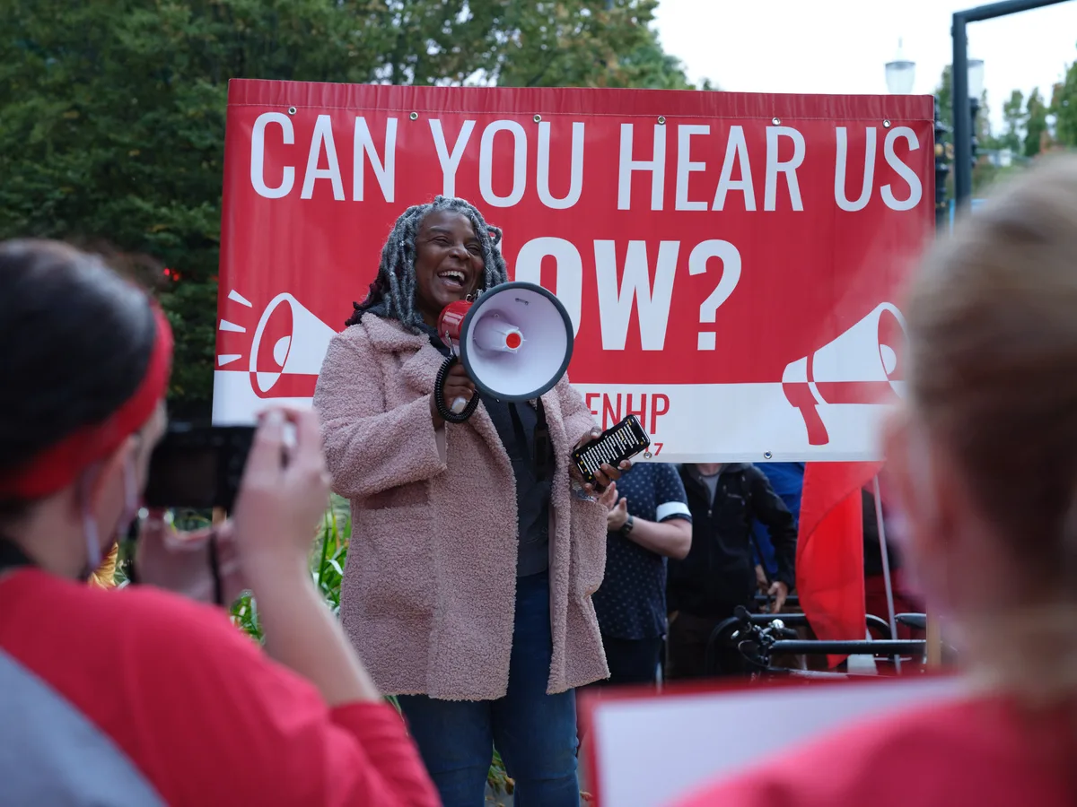 A determined healthcare worker with dreadlocks commands attention through a red and white megaphone, her infectious smile contrasting with the serious nature of the Kaiser Permanente nurses' strike in Portland's Lloyd District. Behind her, a bold crimson banner emblazoned with "CAN YOU HEAR US NOW?" amplifies her message to the sea of red-clad supporters gathered in the foreground. The late afternoon light filters through verdant trees, casting a warm glow over this moment of collective action outside the towering Kaiser Permanente headquarters on Northeast Multnomah Street.