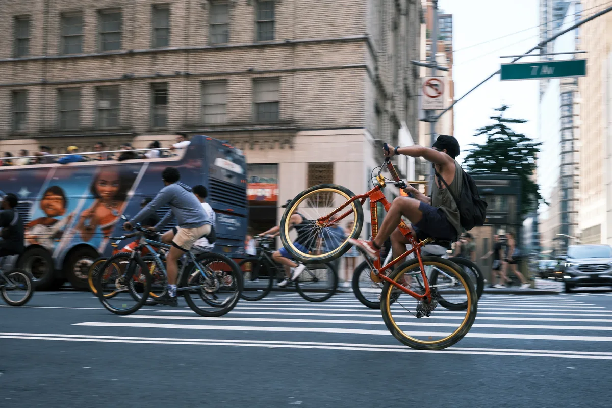 A young cyclist defies gravity, performing a dramatic wheelie on his orange BMX bike as he crosses a busy Manhattan intersection. The motion-blurred scene captures the raw energy of urban street culture, with a city bus and fellow riders creating a dynamic backdrop against the towering brick facades of Midtown. Golden hour light bathes the asphalt, transforming an ordinary crosswalk into a stage for spontaneous street performance.
