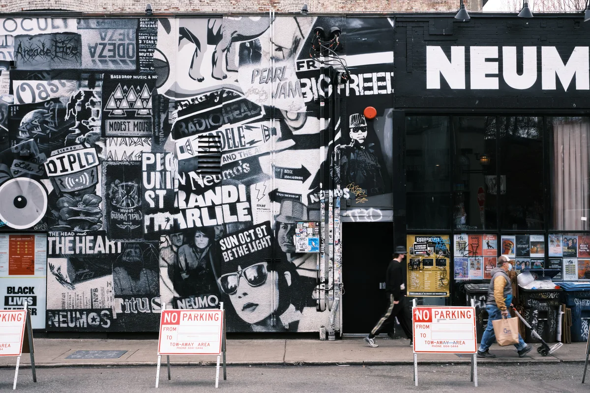 A weathered brick wall in Seattle's Capitol Hill neighborhood serves as a sprawling canvas for layered street art and concert posters, creating a dense collage of black, white, and red imagery. The anarchic composition includes band names like Modest Mouse and Neumos, political messages, and wheat-paste portraits that speak to the district's countercultural identity. Two pedestrians navigate the sidewalk past the artistic chaos, while red 'No Parking' signs punctuate the urban tableau with bureaucratic precision.