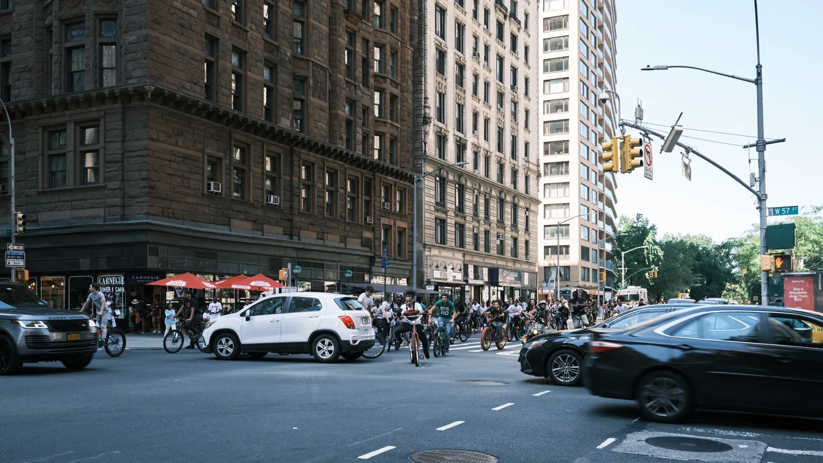 A sea of bicycles clusters at the intersection of West 57th Street and Seventh Avenue in Midtown Manhattan, where riders pause amid the urban choreography of yellow traffic lights and pre-war architecture. The warm afternoon light bathes the weathered brownstone facades and Carnegie Diner's red umbrellas, while modern vehicles blur past in the foreground. This moment captures the tension between old and new New York, where cycling culture claims its space among the towering monuments to commerce and history.