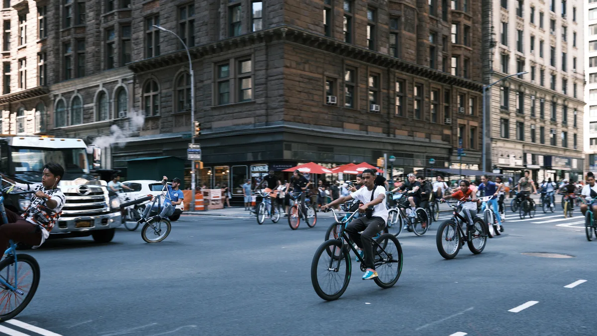 A dynamic street scene captures dozens of cyclists flowing through the intersection at 7th Avenue in Manhattan's Garment District. The afternoon light casts long shadows across the asphalt as riders on various bicycles navigate between yellow cabs and commercial vehicles. Historic stone facades with ornate cornices frame the urban tableau, while red umbrellas from a sidewalk vendor add splashes of color to the predominantly neutral palette of weathered limestone and concrete.