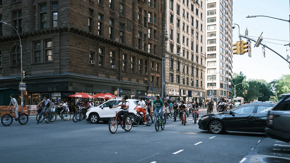 A vibrant street scene captures dozens of cyclists congregating on 7th Avenue in Manhattan, creating an impromptu gathering that transforms the urban thoroughfare into a celebration of bicycle culture. The warm afternoon light bathes the historic brownstone facades and creates long shadows across the asphalt, while modern vehicles navigate around the cycling enthusiasts. The composition juxtaposes New York's architectural heritage—from weathered sandstone buildings to contemporary high-rises—with the grassroots energy of the city's cycling community.