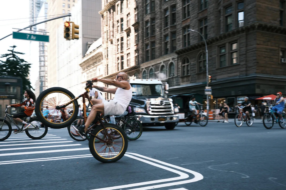 A shirtless cyclist in white shorts performs a dramatic wheelie on his custom bicycle, commanding the center of 7th Avenue in Midtown Manhattan. The golden hour light bathes the historic architecture and creates dynamic shadows across the bustling intersection, while traffic and pedestrians blur into the urban rhythm. The scene captures the raw energy and improvisational spirit of New York street culture against the towering backdrop of Manhattan's iconic skyline.
