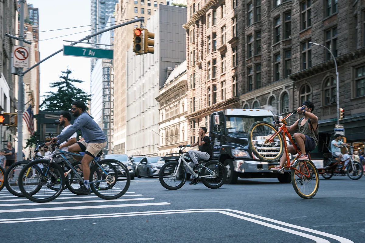 A dynamic street scene captures the rhythm of Manhattan transportation as cyclists of varying ages navigate the intersection at 7th Avenue. Golden hour light bathes the historic stone facades and modern glass towers, creating dramatic contrast between the pre-war architecture and contemporary urban mobility. The composition frames multiple cyclists mid-motion against the backdrop of Midtown's architectural tapestry, while pedestrians and traffic create layers of metropolitan energy.