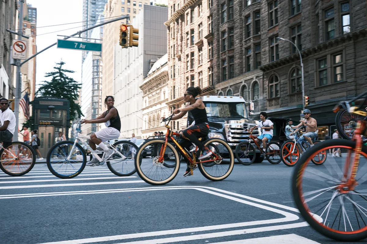 A dynamic street scene captures cyclists in motion along 7th Avenue in Manhattan's Midtown, their wheels blurred against the crisp asphalt crosswalk stripes. The golden hour light bathes the historic stone facades and modern glass towers that define this iconic corridor, while pedestrians and vehicles share the urban rhythm. The composition emphasizes the democratic nature of city streets, where bicycles weave through the metropolitan tapestry alongside cars and buses.