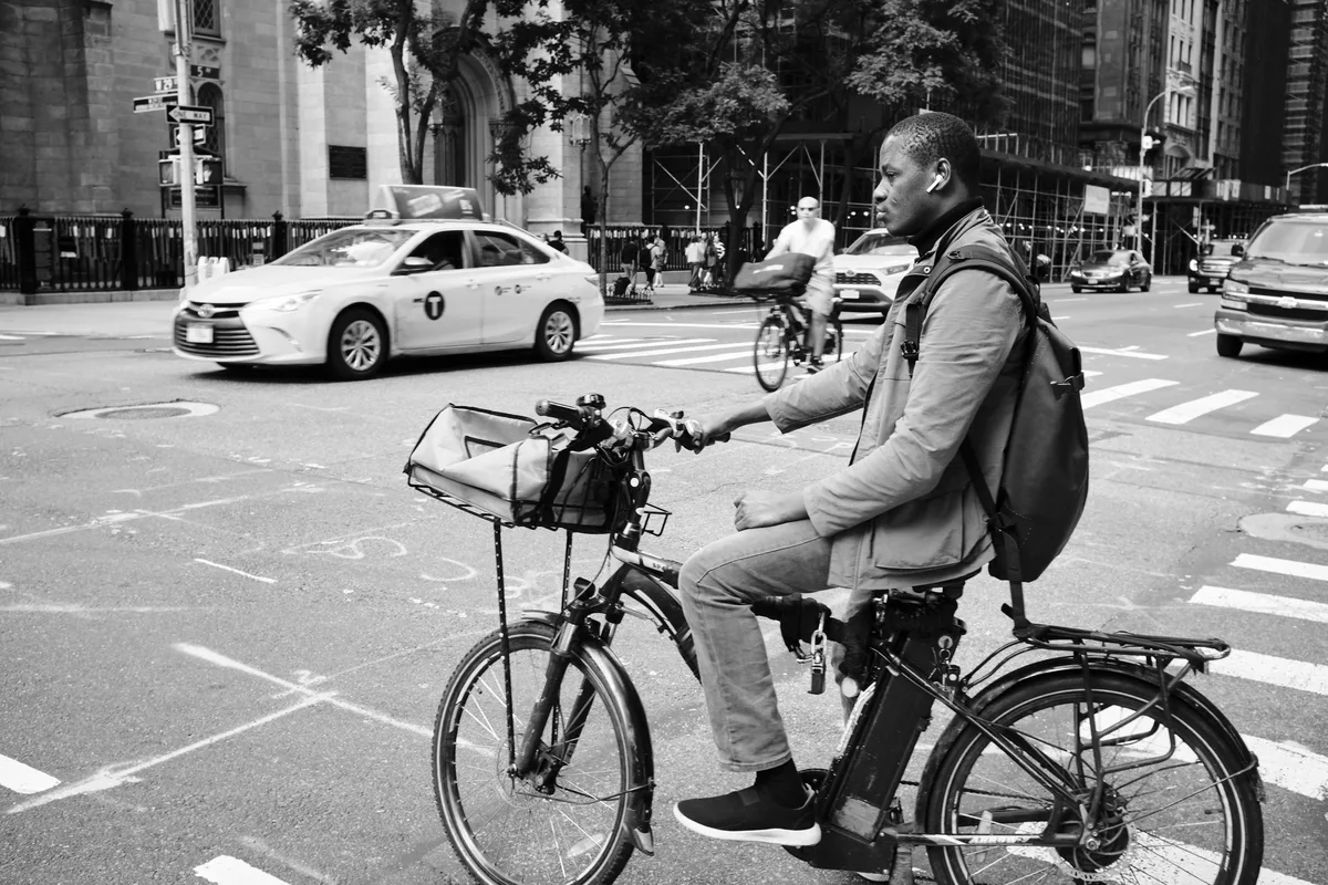 A delivery cyclist pauses at the intersection of East 29th Street in Manhattan's NoMad district, his weathered face caught in contemplative profile against the bustling urban backdrop. The black and white composition captures the gritty authenticity of city life, with his utilitarian bicycle equipped with a front cargo basket positioned prominently in the crosswalk. Behind him, the familiar yellow taxi and Gothic Revival architecture of the neighborhood create a quintessentially New York tableau, while dappled sunlight filters through street trees onto the asphalt.