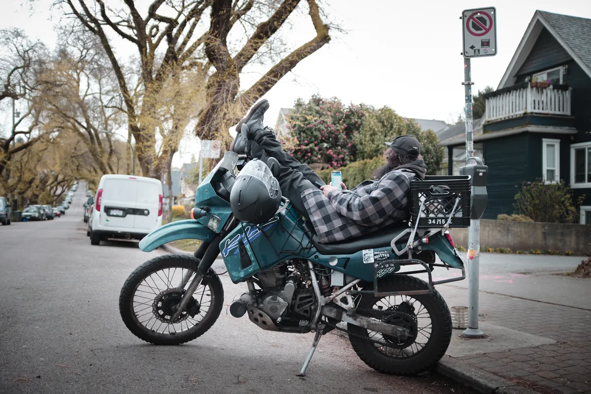 A weathered adventure motorcycle loaded with travel gear sits parked on East 6th Avenue in Vancouver's Grandview-Woodland neighborhood, its rider taking a contemplative break. The teal dual-sport bike, adorned with scratches and travel stickers, bears witness to countless miles beneath the bare branches of winter trees. Residential charm surrounds the scene with heritage homes and urban signage creating an atmospheric backdrop under overcast skies.
