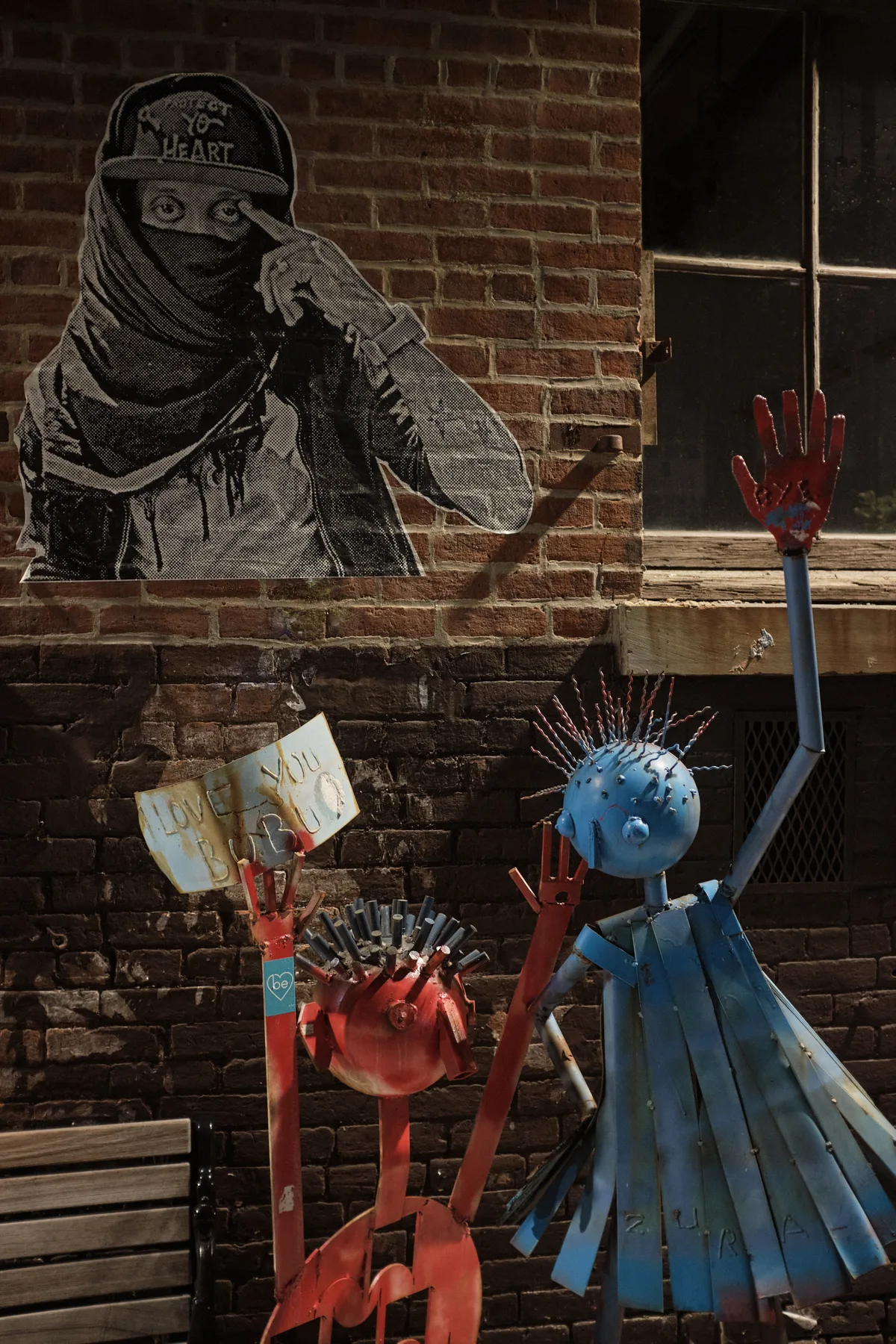 A provocative street art tableau unfolds against weathered brick in Brooklyn's Williamsburg neighborhood, where anonymous rebellion meets whimsical protest. A masked figure rendered in stark black and white wheat paste dominates the wall, bearing the cryptic message 'NY HEART,' while below, two vibrant sculptural figures—one crimson, one azure—raise defiant hands skyward in a gesture of solidarity. The afternoon light catches the textured surfaces and peeling paint, creating a dialogue between the ephemeral nature of street art and the permanent urban landscape.