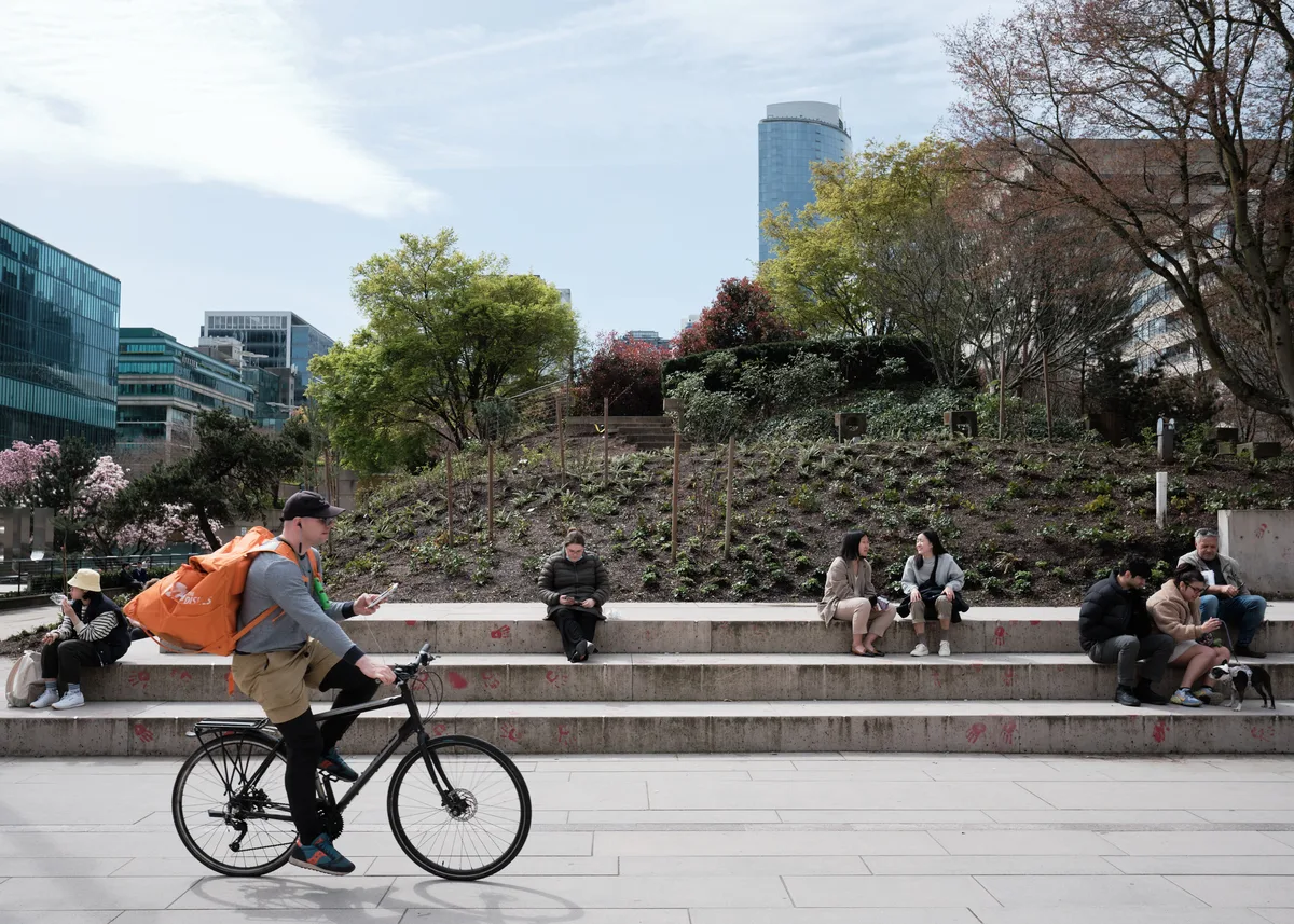 A delivery cyclist in a vibrant orange jacket glides past tiered concrete steps where young people gather in casual clusters beneath spring's awakening foliage. The scene unfolds against Vancouver's distinctive downtown skyline, with glass towers rising behind a hillside garden showing the tender greens and burgundy blossoms of the Pacific Northwest's seasonal transition. Soft overcast light bathes the urban plaza, creating an intimate tableau of city life in motion.