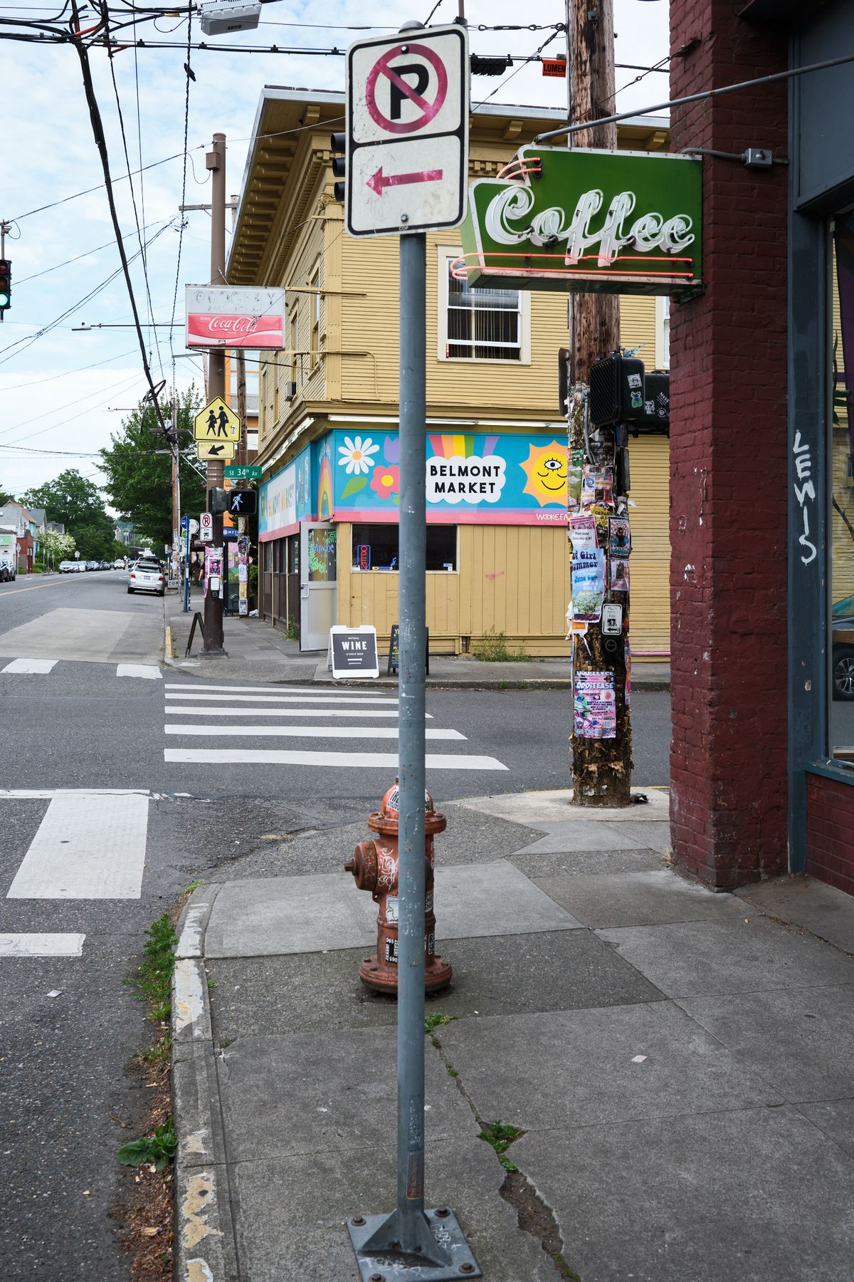 A street corner view in Portland, Oregon showing a crosswalk, no right turn sign, fire hydrant, and local businesses including Belmont Market and a coffee shop. The scene captures the eclectic urban character of the Belmont district with colorful storefronts, utility poles, and mixed-use buildings.