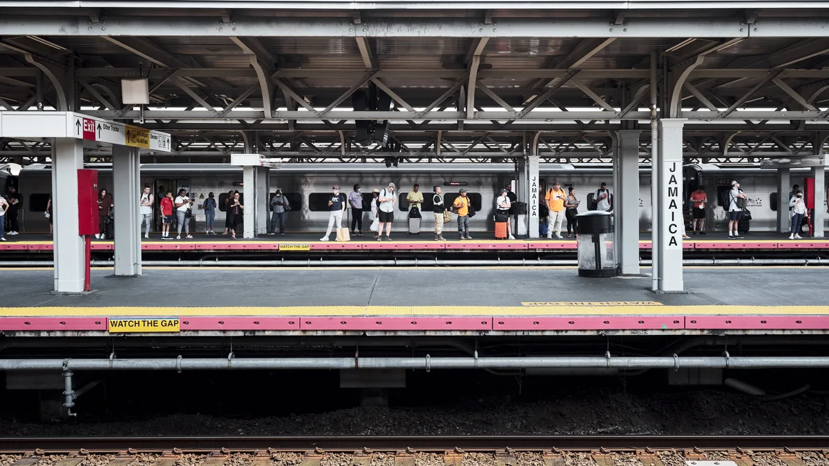 Passengers disperse across the weathered platform of Jamaica Station, their colorful summer clothing creating vibrant punctuation marks against the industrial backdrop of steel beams and concrete pillars. The AirTrain car sits silent between destinations, its sleek silver form contrasting with the utilitarian architecture of this crucial transportation hub. Harsh fluorescent lighting mingles with natural daylight filtering through the elevated structure, casting complex shadows that emphasize the geometric patterns of the overhead framework.