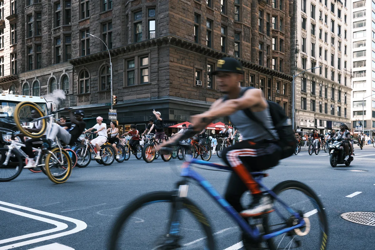 Motion blur captures the kinetic energy of dozens of cyclists flowing down 7th Avenue in Midtown Manhattan, their forms ghosted against the solid backdrop of historic brownstone architecture. The foreground rider in a blue cap emerges sharp from the stream of blurred movement, while golden-rimmed bicycle wheels catch fragments of afternoon light. Behind the cycling parade, the stately Carnegie Hall neighborhood's characteristic sandstone facades provide timeless contrast to this modern urban ballet of sustainable transportation.