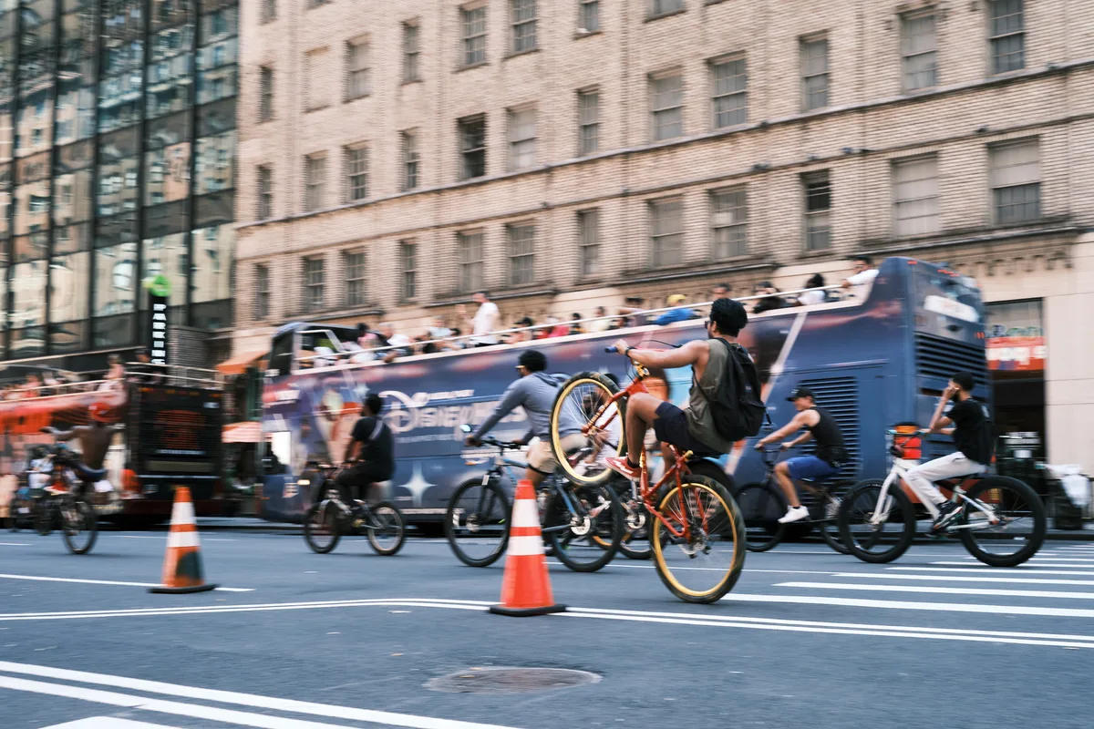 Motion blur captures the kinetic energy of cyclists racing down Manhattan's Seventh Avenue, their figures streaking past in a symphony of movement against the stoic backdrop of brick tenements and modern glass facades. The photographer's intentional camera movement transforms the urban thoroughfare into a river of color and speed, where orange traffic cones punctuate the asphalt like sentinels guiding the flow of human-powered transit. The contrast between the sharp architectural details and the fluid cyclists creates a visceral sense of metropolitan rhythm and the relentless pulse of New York City street life.