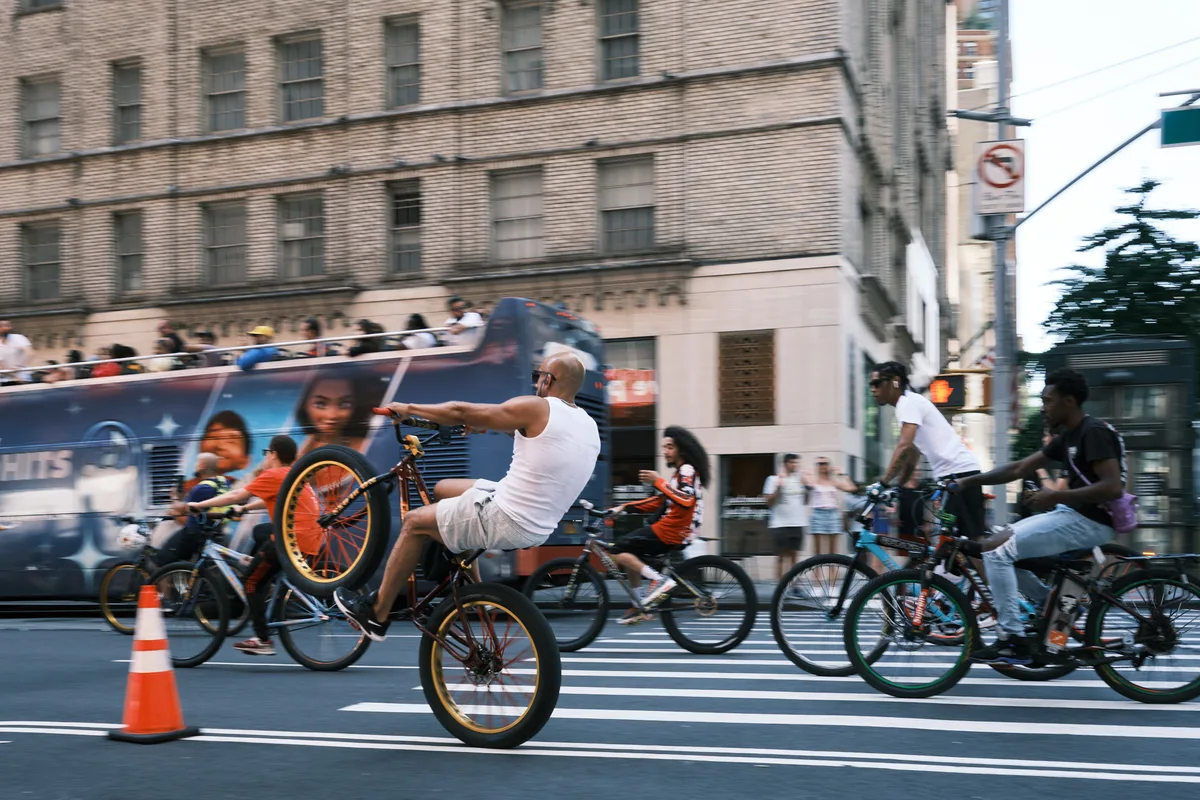 A dynamic street scene captures the kinetic energy of Manhattan's cycling culture as riders navigate the crosswalk at 7th Avenue. The central figure, a bald man in a white tank top, executes a dramatic wheelie on his bicycle with orange-rimmed wheels, his body language expressing pure urban swagger. Behind him, a blue city bus creates a striking backdrop against the weathered brick facades of Midtown buildings, while other cyclists and pedestrians blur through the frame, suggesting the constant motion of New York street life. The golden hour light bathes the scene in warm tones, transforming an ordinary intersection into a stage for spontaneous urban theater.