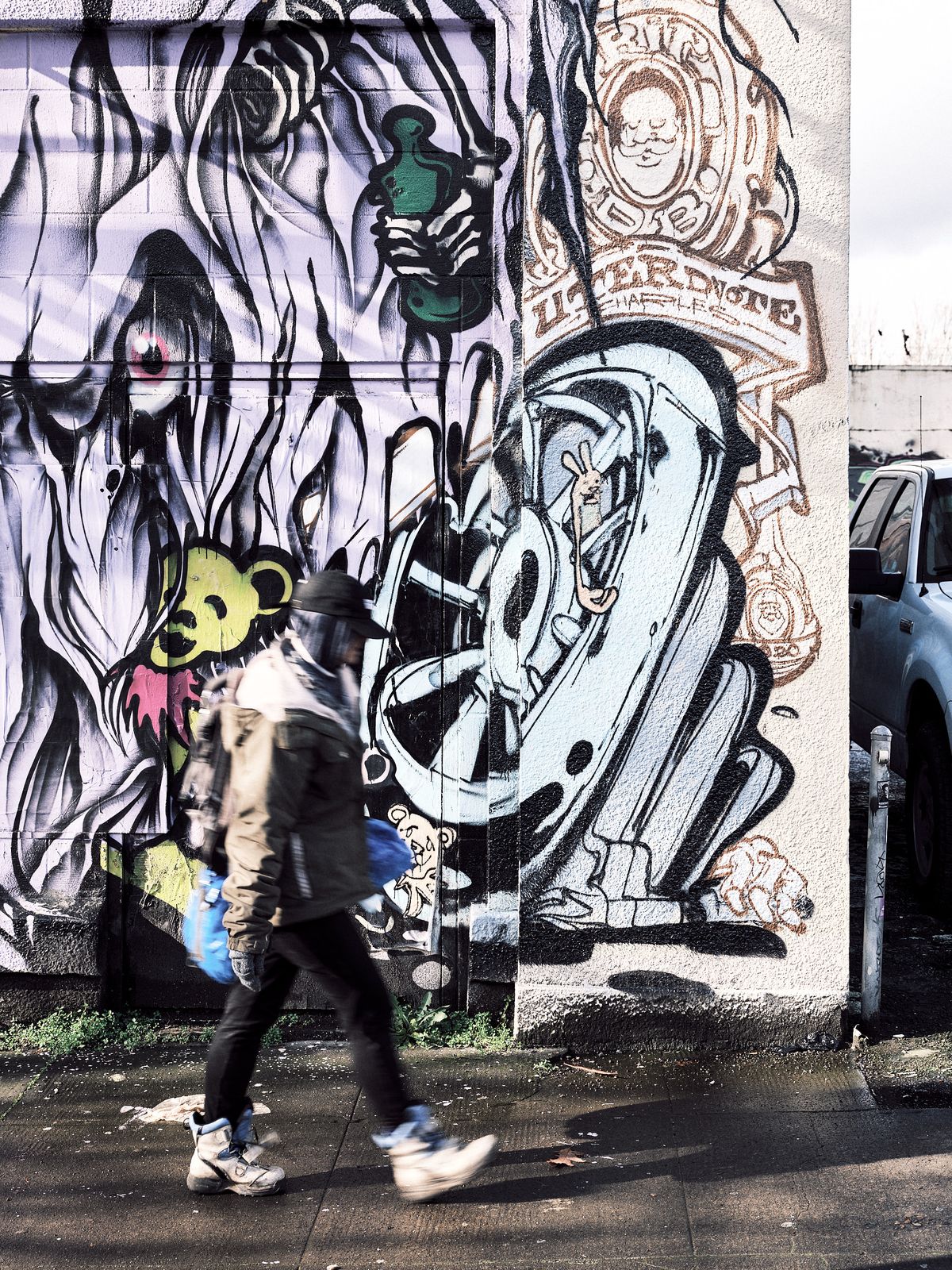 A motion-blurred pedestrian carrying a blue bag walks briskly along a wet sidewalk in Portland, Oregon, past a large-scale graffiti mural by artist Uter. The mural spans two building surfaces and features bold black-and-white surrealist imagery including a hooded figure, a cartoon bear character in yellow and pink, a stylized letter form with a rabbit inside, and circular emblems with illustrated faces. A parked white truck is partially visible on the right.