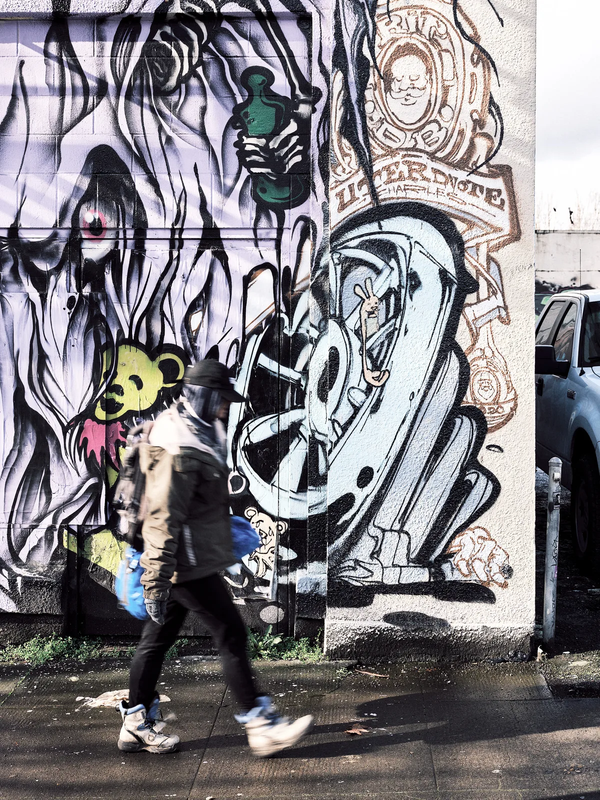 A motion-blurred pedestrian carrying a blue bag walks briskly along a wet sidewalk in Portland, Oregon, past a large-scale graffiti mural by artist Uter. The mural spans two building surfaces and features bold black-and-white surrealist imagery including a hooded figure, a cartoon bear character in yellow and pink, a stylized letter form with a rabbit inside, and circular emblems with illustrated faces. A parked white truck is partially visible on the right.
