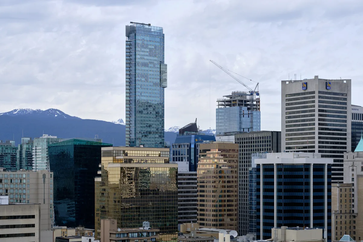 The gleaming downtown core of Vancouver rises in sharp architectural contrast against the snow-dusted North Shore Mountains. A commanding blue glass tower dominates the skyline while construction cranes hint at the city's relentless growth. The overcast Pacific Northwest sky creates a muted palette that emphasizes the interplay between urban ambition and natural grandeur, with the coastal range providing a dramatic backdrop to this dense collection of office buildings and residential towers.