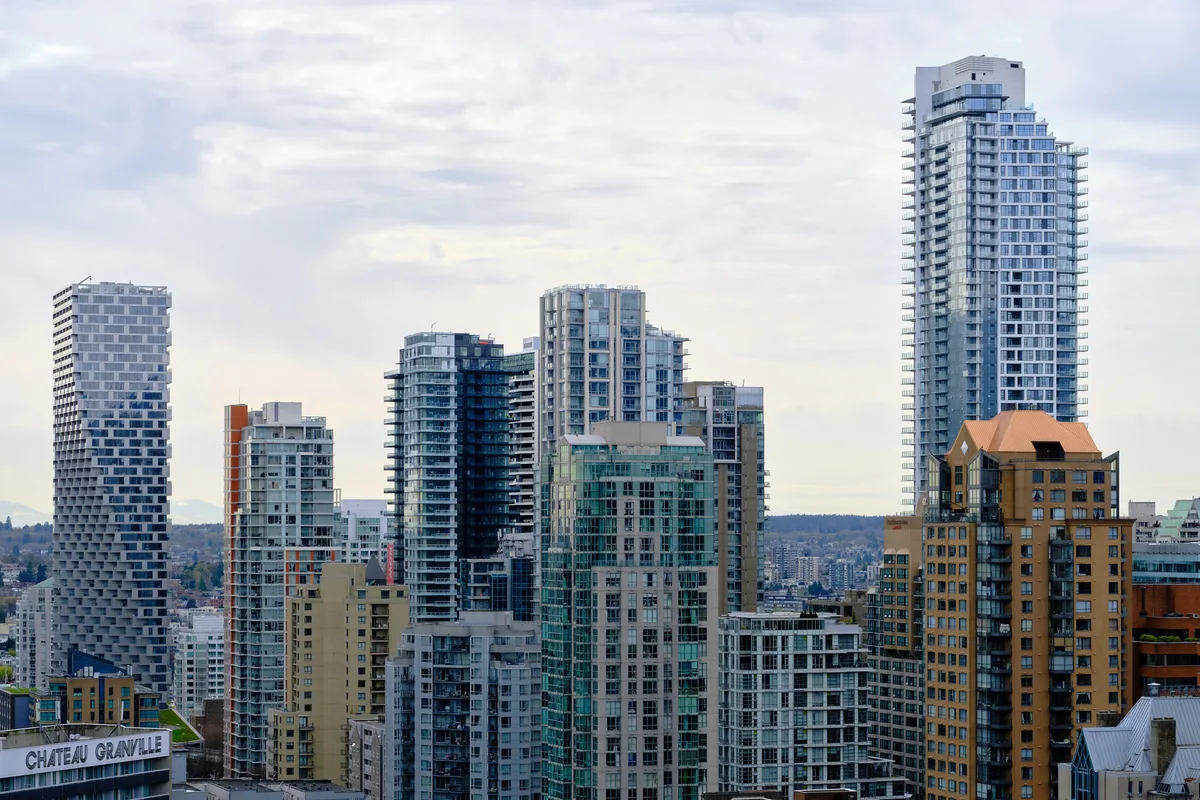 A dense cluster of contemporary residential towers rises against Vancouver's moody Pacific Northwest sky, their glass and concrete facades catching the diffused light of an overcast afternoon. The architectural composition showcases the city's modern urban density, with sleek high-rises in blues, grays, and warm terracotta tones creating a striking vertical landscape. In the foreground, the historic Chateau Granville sign anchors the scene, while forested hills stretch toward the horizon, emphasizing Vancouver's unique blend of urban sophistication and natural beauty.
