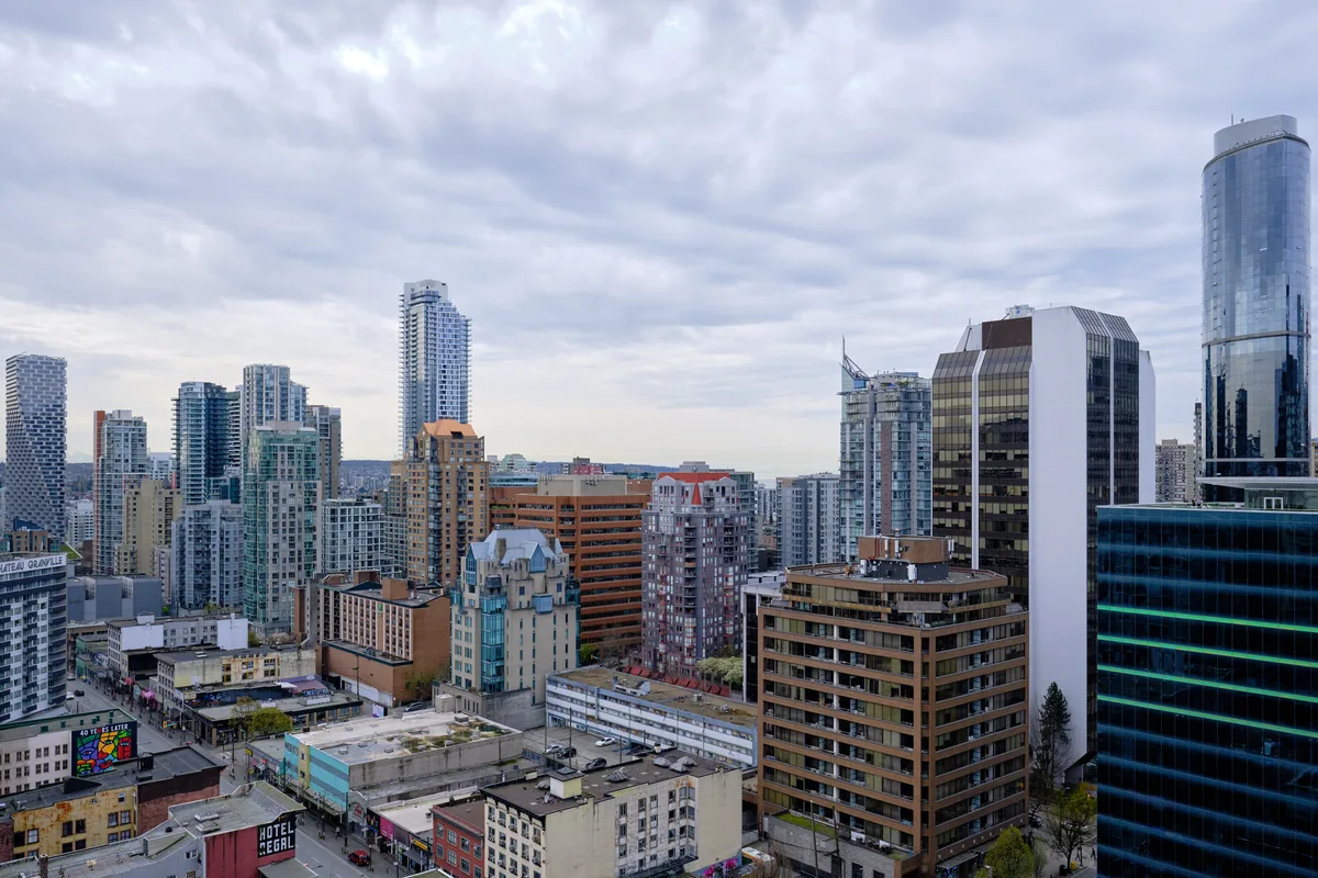 A sweeping aerial view of Vancouver's dense downtown core captured beneath a dramatic canopy of pewter clouds. The urban landscape unfolds in layers of architectural diversity, from gleaming glass towers to weathered brick facades, creating a complex metropolitan mosaic. Muted daylight filters through the overcast sky, casting the city in subdued tones that emphasize the geometric interplay between old and new structures. The composition reveals the organic density of urban development, where residential high-rises, commercial buildings, and street-level establishments converge in Vancouver's characteristic west coast urbanism.