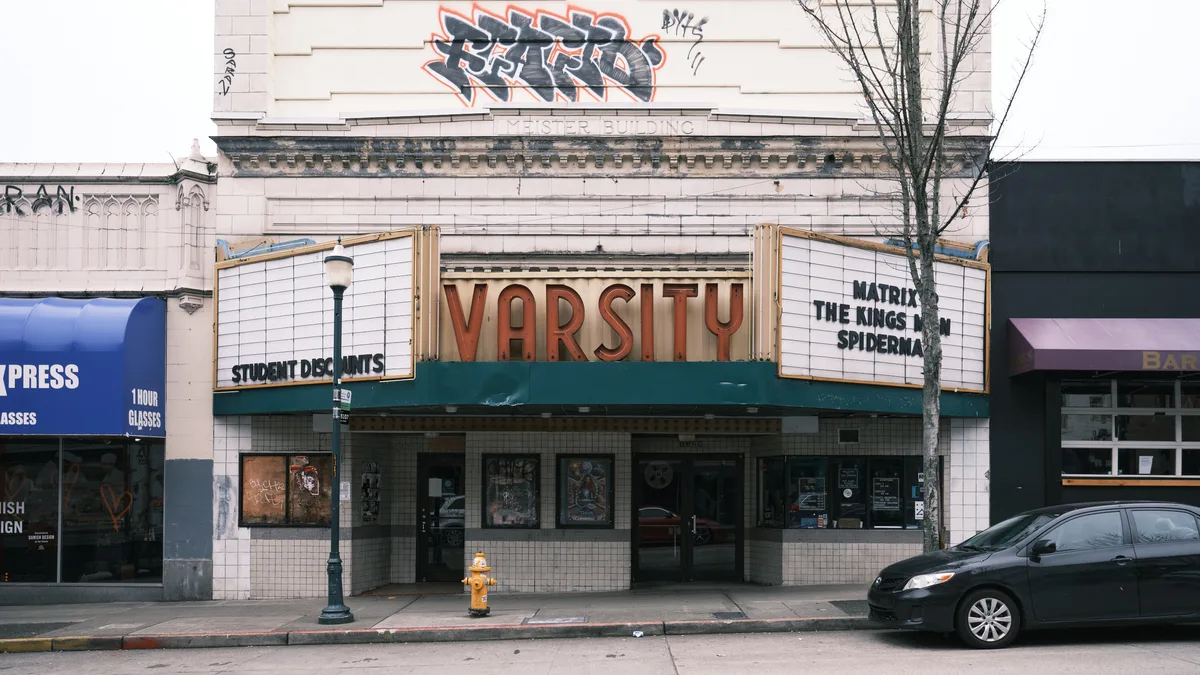 The historic Varsity Theatre stands sentinel along University Way Northeast in Seattle's University District, its weathered white brick facade crowned with bold graffiti art. The classic movie theater's red marquee letters spell out "VARSITY" above a green canopy, while the adjacent signage advertises "STUDENT DISCOUNTS" and current films including "MATRIX" and "THE KING SPIDERMAN." Overcast daylight illuminates this quintessential college town streetscape, where bare winter branches frame the vintage cinema amid neighboring storefronts and parked cars.