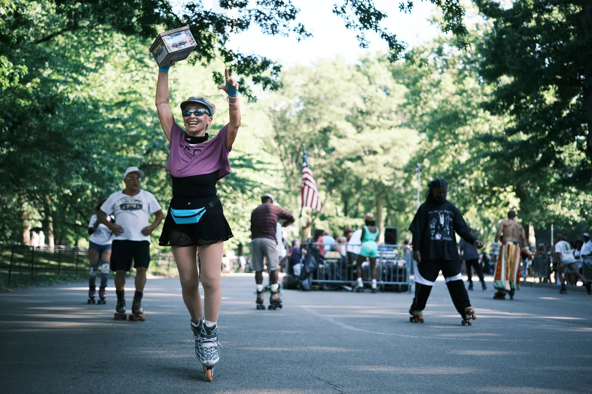 A triumphant roller skater in purple athletic wear raises both arms in celebration while gliding down Central Park's tree-lined pathway, a trophy box held high above her head. The scene captures pure joy as filtered sunlight creates dancing patterns through the dense summer canopy, while fellow skaters and park visitors populate the background beneath an American flag. The composition draws the eye to her moment of victory against the park's iconic verdant tunnel, creating an atmosphere of community celebration and athletic achievement.