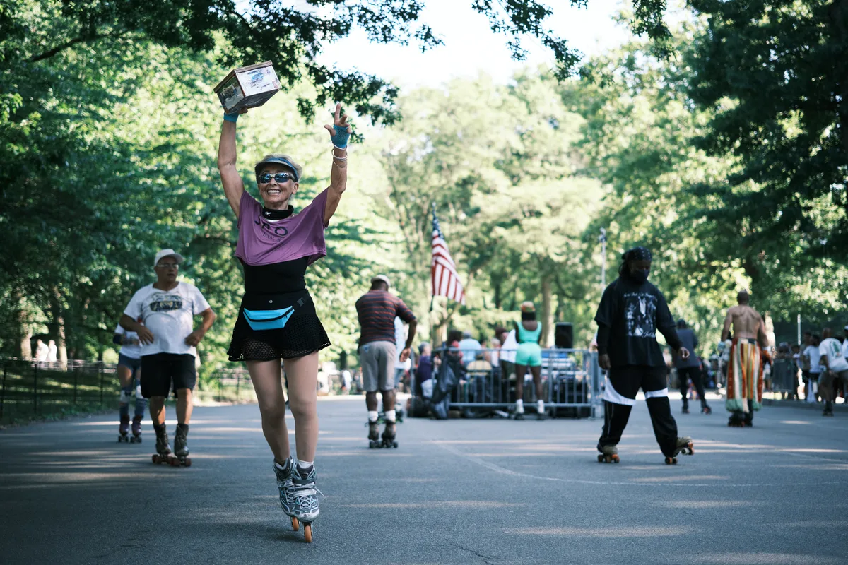 A jubilant roller skater in purple and black athletic wear celebrates triumphantly on Central Park's tree-lined pathways, both arms raised high while clutching what appears to be an award or trophy. Dappled sunlight filters through the dense summer canopy, creating a luminous backdrop as other skaters glide leisurely in the distance. The American flag stands proudly among the verdant trees, while the smooth asphalt stretches invitingly ahead, capturing the pure joy of recreational skating in Manhattan's beloved green heart.