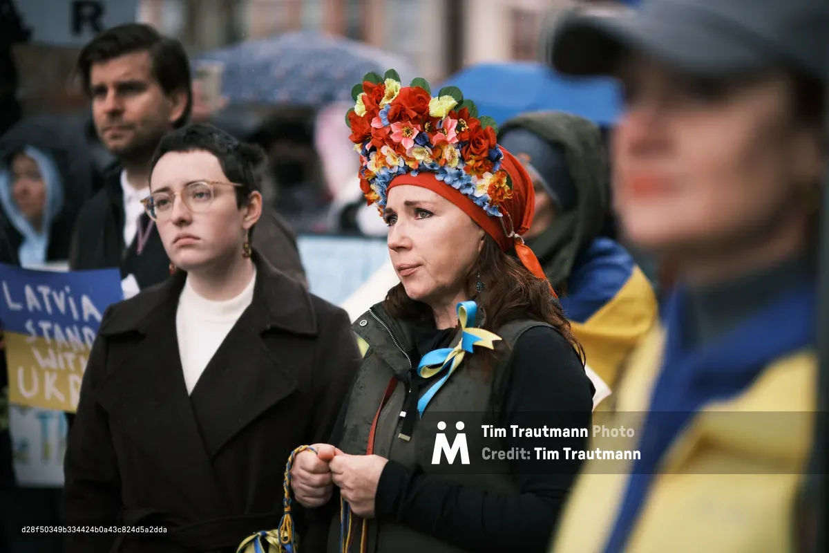 Two women stand in somber solidarity outside Portland's Revolution Hall, their faces etched with quiet determination as they participate in an anti-war demonstration days before Russian hostilities would begin in Ukraine. The woman on the right wears a traditional Ukrainian floral headdress crowned with vibrant red and blue blooms, while blue and yellow flags flutter in the background like whispered prayers for peace. The overcast Pacific Northwest light casts a prophetic shadow over this prescient gathering, capturing a moment suspended between hope and inevitable tragedy.
