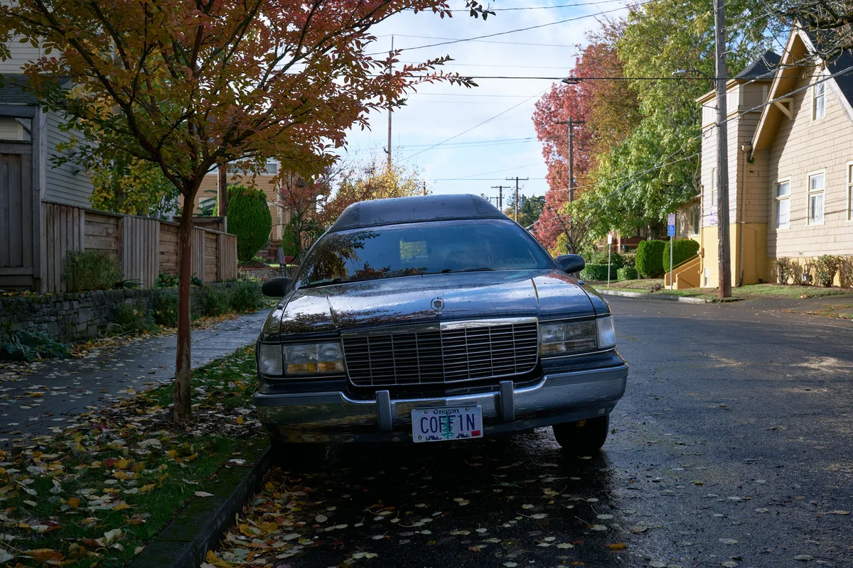 A black vintage hearse with Oregon license plate 'COFFIN' is parked on a wet asphalt street in a residential Portland neighborhood during autumn. The scene features colorful fall foliage, wooden fences, and characteristic Pacific Northwest housing architecture.